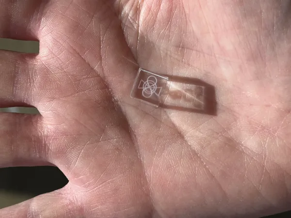 A photo of a palm of a hand, that has a small glass cube with some engraved concentric forms: an eight, a circle and two triangles
